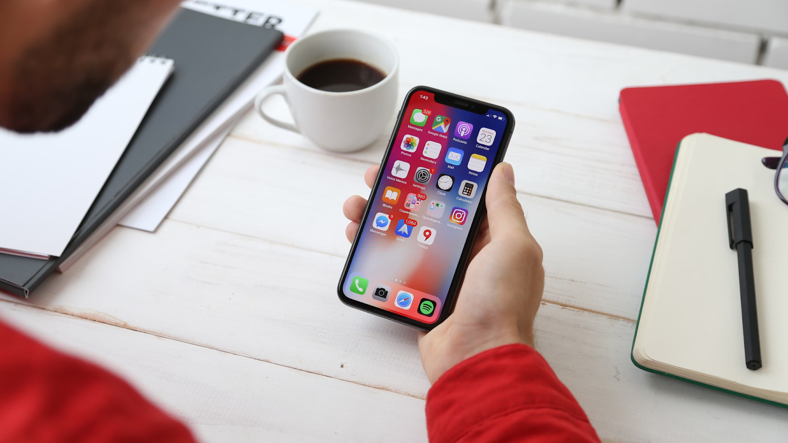 Person holding a smartphone with various apps open on the home screen, seated at a white desk with a coffee cup, notebooks, and pen