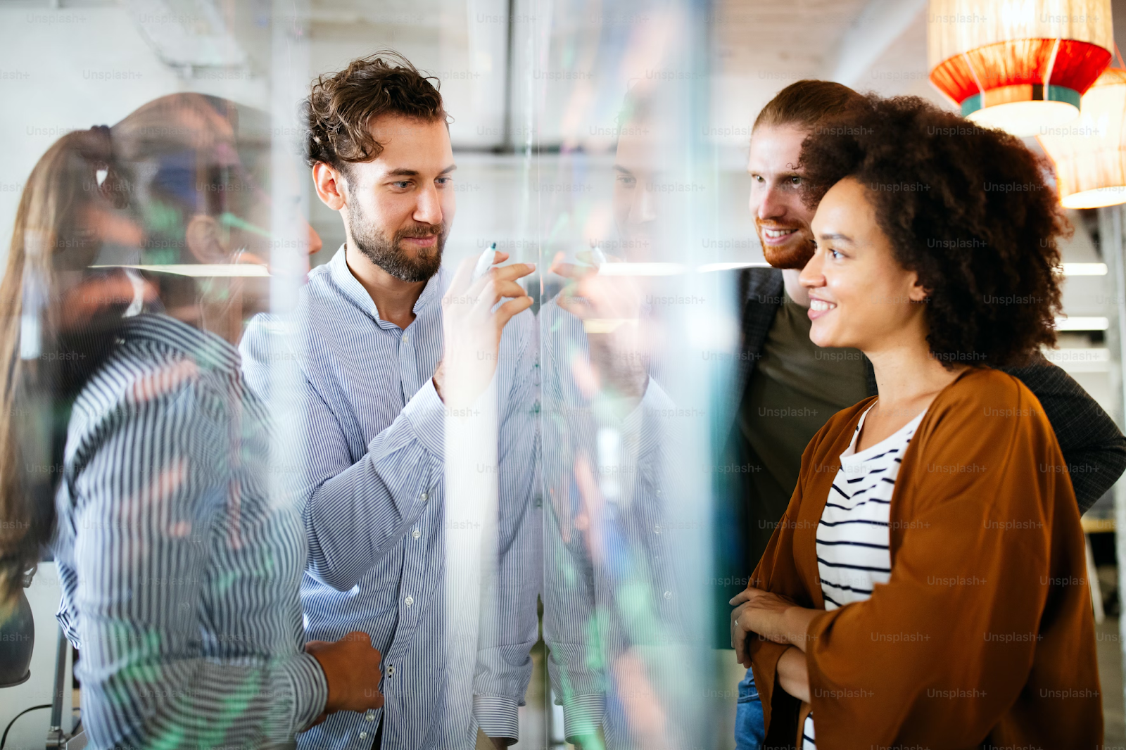 Team of diverse professionals collaborating at a glass board in a modern office, brainstorming ideas and discussing strategies, with reflections adding a creative visual effect.